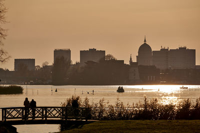 Silhouette buildings by river against sky during sunset