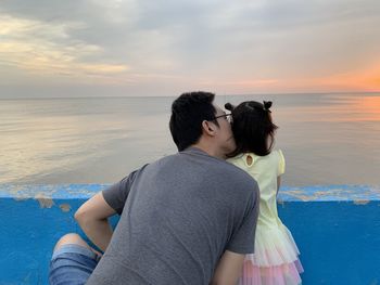 Rear view of father kissing daughter against sea during sunset