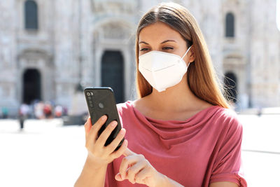 Close-up of young woman wearing flu mask using smart phone outdoors