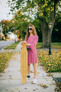 Girl wearing sunglasses standing on sidewalk holding a skateboard