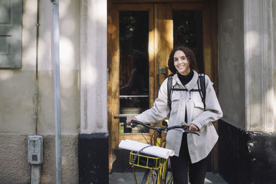 Portrait of smiling female architect standing with bicycle at building entrance