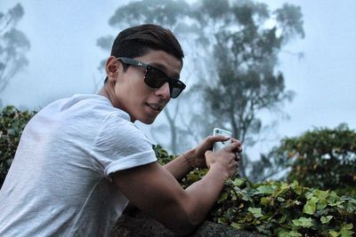 Portrait of young man wearing sunglasses holding mobile phone while standing by plants in park during foggy weather