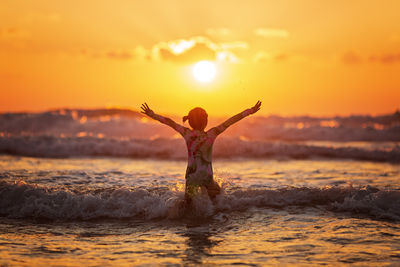 Rear view of woman with arms raised at beach against sky during sunset