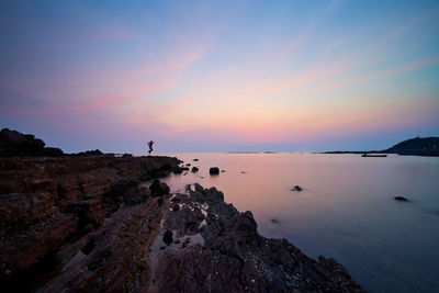 Mid distance view of person jumping on rock formation by sea at sunset