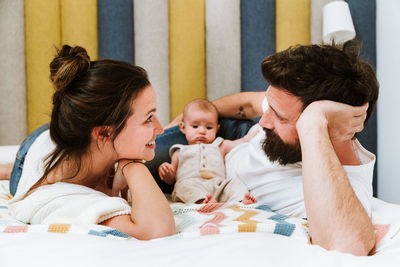 Playful father playing with baby while chilling together with wife and kid on soft bed at home looking at each other
