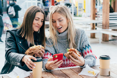 Two women eat fast food, drink coffee and watch social media while sitting at a table outside.