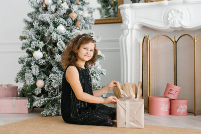 A seven-year-old girl in a black dress with sequins sits near the fireplace at the christmas tree