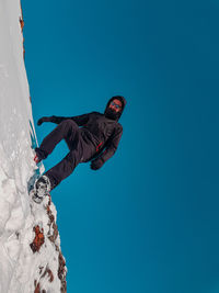 Low angle view of young man standing on snow covered mountain against clear blue sky