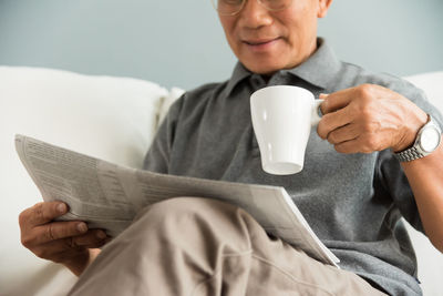Midsection of smiling senior man with tea holding newspaper while sitting on sofa