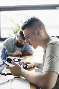 Male computer programmer using smart phone by laptop on desk in startup company