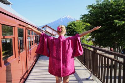 Rear view of woman with arms raised standing at railroad station platform against clear sky