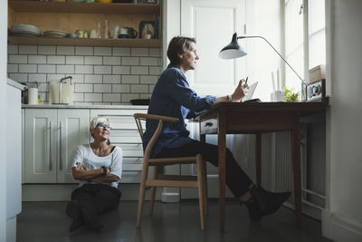 Female industrial designer looking at colleague while sitting on floor in kitchen
