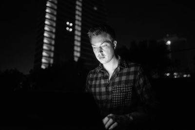 Portrait of young man looking away while sitting in city at night