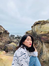 Portrait of woman sitting on rock at beach against cloudy sky