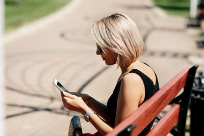 A young woman takes a selfie sitting on a park bench, adjusts her makeup and hairstyle