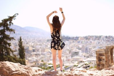 Rear view of young woman with arms raised standing on rock against cityscape
