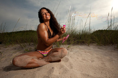 Full length of woman with bubble wand sitting on sand against sky