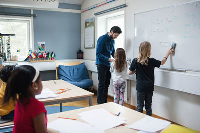 Girl talking to teacher while student drawing on whiteboard in classroom