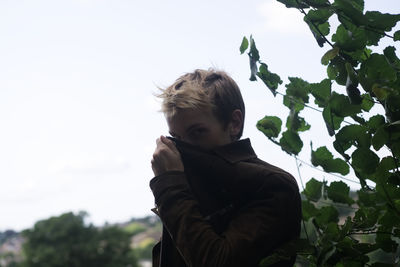 Low angle portrait of boy standing by tree against sky