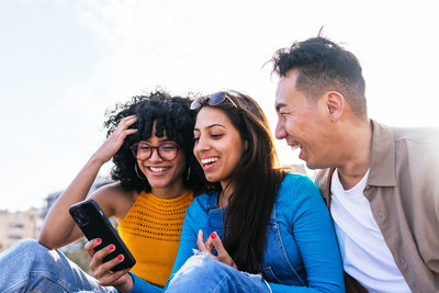 From below happy multiracial man and women smiling and browsing social media on cellphones while resting on beach on sunny summer day on resort