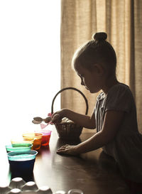 Side view of girl preparing easter eggs on table by window at home