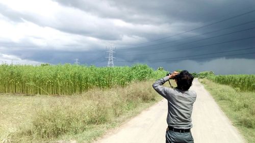 Rear view of man photographing farm and electricity pylon against cloudy sky