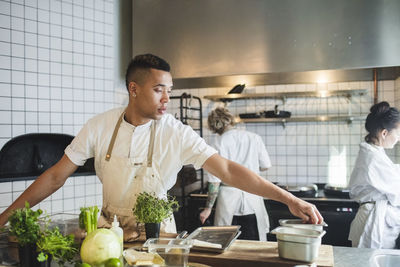 Young male chef cooking in kitchen against colleagues working at restaurant