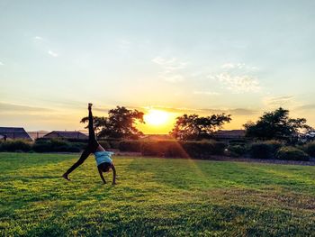 Rear view of girl doing cartwheel on grassy field at park against sky during sunset