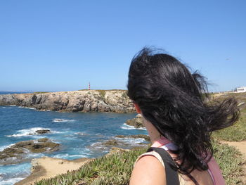 Woman looking at lighthouse on cliff at sea shore against clear blue sky