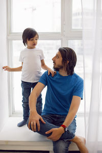 Father in a blue t-shirt and a son in a white t-shirt. sitting on the windowsill of a large window 