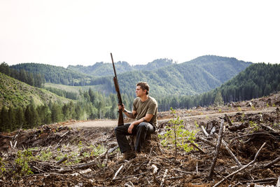 Full length of hunter sitting on tree stump against mountains