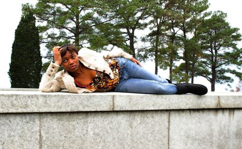 Thoughtful woman lying on retaining wall against trees