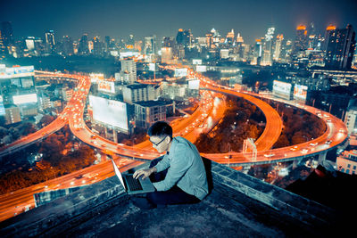 Man sitting at illuminated cityscape against sky at night