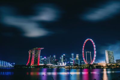 Illuminated ferris wheel by city against sky at night