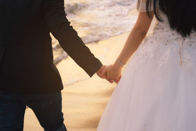 Midsection of bride and bridegroom holding hands on shore at beach