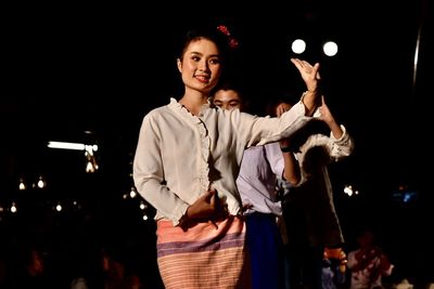 Portrait of young woman with arms raised standing at night