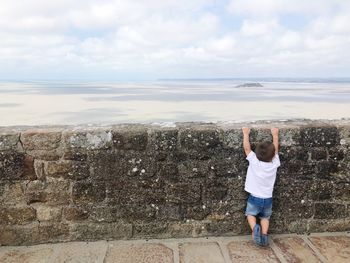 Rear view of boy hanging on retaining wall against sea