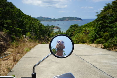 Reflection of man on side-view mirror by sea against sky