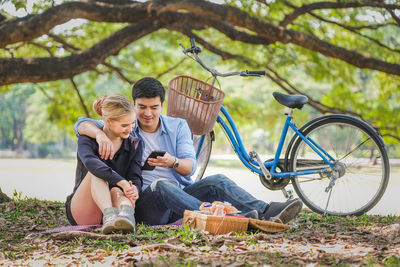 Man using mobile phone while sitting with woman on grass by bicycle