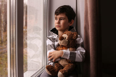 The boy dreamily looks out the window, hugging a teddy bear