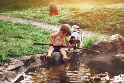 Full length of shirtless boy holding stick sitting by pond with dog