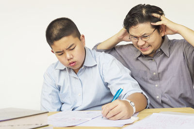 Frustrated father teaching son while sitting at table against white background