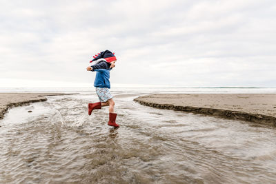 Young child wearing knit dinosaur hat jumping at beach