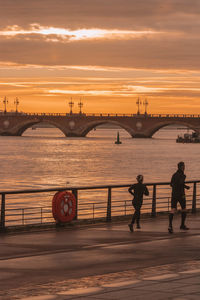 People on bridge over river against sky during sunset