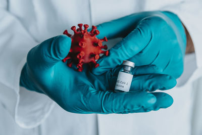 Close-up of scientist hand holding vaccine bottle and coronavirus model while standing at laboratory