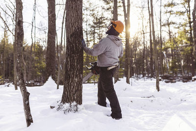 Side view of lumberjack holding chainsaw while standing by tree trunk in forest during winter