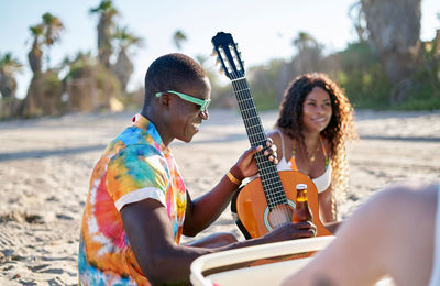 African american male musician in stylish colorful shirt and sunglasses drinking beer while sitting on sandy beach with acoustic guitar and talking to girlfriend during summer vacation