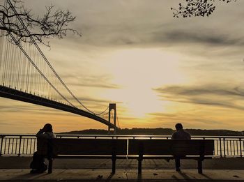 Man and woman sitting on bench against suspension bridge over river