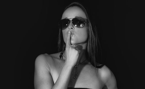 Close-up portrait of woman in sunglasses with finger on lips against black background