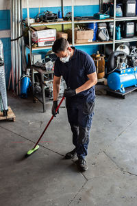 High angle of man in uniform and respirator sweeping floor with broom while working in garage